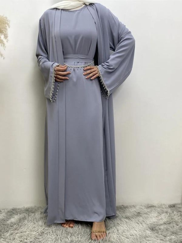 Femme en abaya bleu ciel avec des broderies argentées aux manches et à la taille, portant un hijab blanc, posant avec une main sur la hanche, sur un fond neutre avec un vase de pampas à gauche