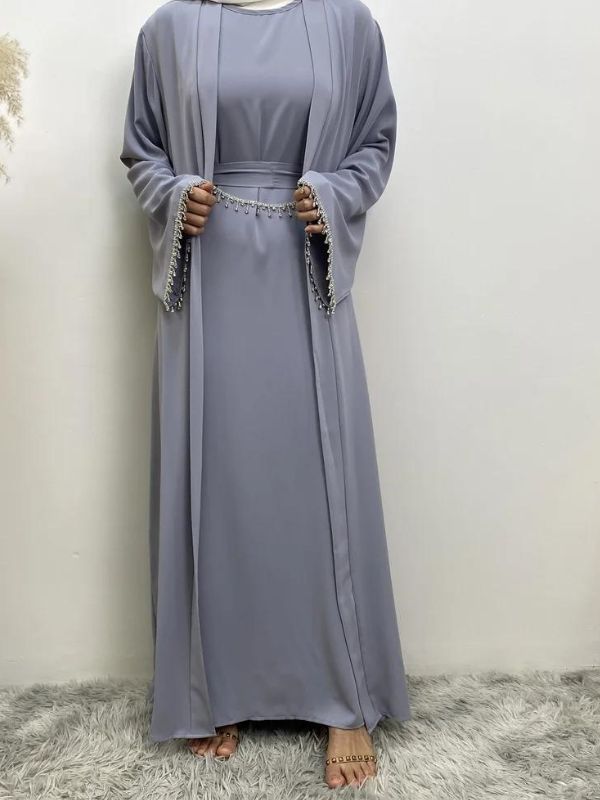 Femme en abaya bleu ciel avec des broderies argentées aux manches et à la taille, portant un hijab blanc, posant avec une main sur la hanche, sur un fond neutre avec un vase de pampas à gauche