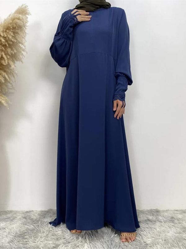 L'image montre une femme portant une abaya longue de couleur bleue, vue de face. La robe est fluide et tombe gracieusement jusqu'au sol. Les manches de la robe sont ornées de détails plissés au niveau des poignets. La femme porte un hijab vert olive qui encadre son visage et couvre ses cheveux. 