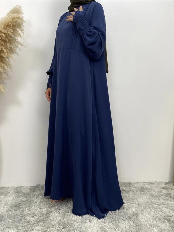 L'image montre une femme portant une abaya longue de couleur bleue, vue de face. La robe est fluide et tombe gracieusement jusqu'au sol. Les manches de la robe sont ornées de détails plissés au niveau des poignets. La femme porte un hijab vert olive qui encadre son visage et couvre ses cheveux. 