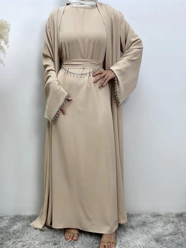 Femme portant une abaya champagne ornée de broderies argentées aux manches et à la taille, portant un hijab blanc, posant avec une main sur la hanche et l'autre près de la taille, sur un fond neutre avec un vase de pampas à gauche