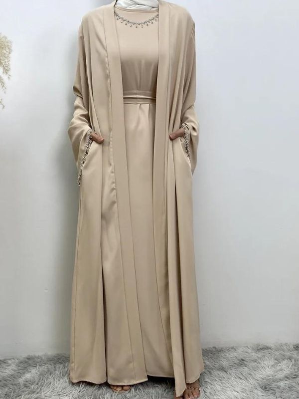 Femme portant une abaya champagne ornée de broderies argentées aux manches et à la taille, portant un hijab blanc, posant avec une main sur la hanche et l'autre près de la taille, sur un fond neutre avec un vase de pampas à gauche