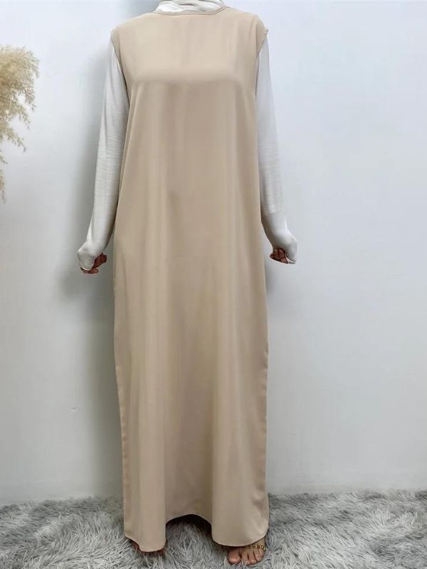 Femme portant une robe abaya champagne par-dessus un haut à manches longues blanc, posant sur un fond neutre avec un vase de pampas à gauche