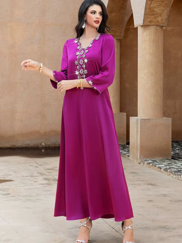 Abaya Dubaï fuchsia, vue de face en pied entre des colonnes, un bras tendu sur le côté et l'autre main devant la poitrine, manches évasées bordées de strass