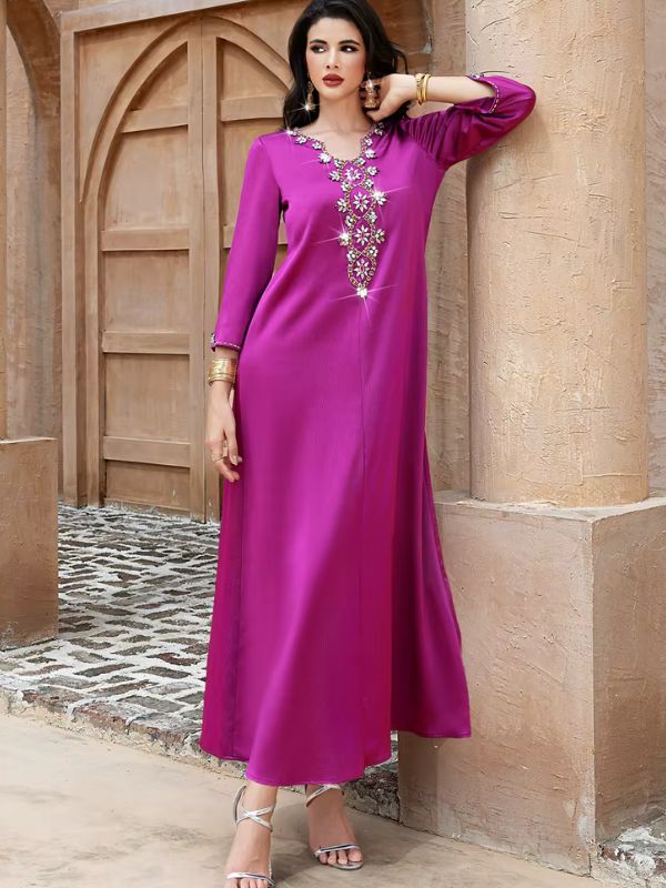 Abaya Dubaï fuchsia, vue de face en pied, appuyée contre un pilier devant une porte en bois, main posée près du cou