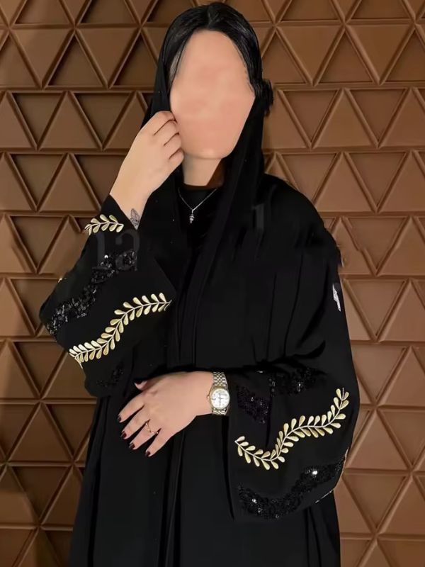 Femme voilée portant une abaya noire aux manches brodées de feuillages dorés, posant contre un mur géométrique
