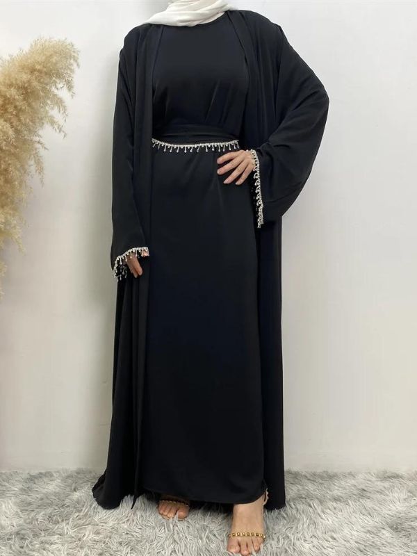 Femme portant une abaya noire avec des détails en broderie blanche aux manches et à la taille, accompagnée d'un hijab blanc, posant avec une main sur la hanche sur un fond neutre