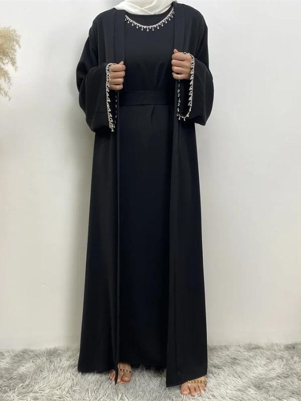 Femme portant une abaya noire avec des détails en broderie blanche aux manches et à la taille, accompagnée d'un hijab blanc, posant avec une main sur la hanche sur un fond neutre