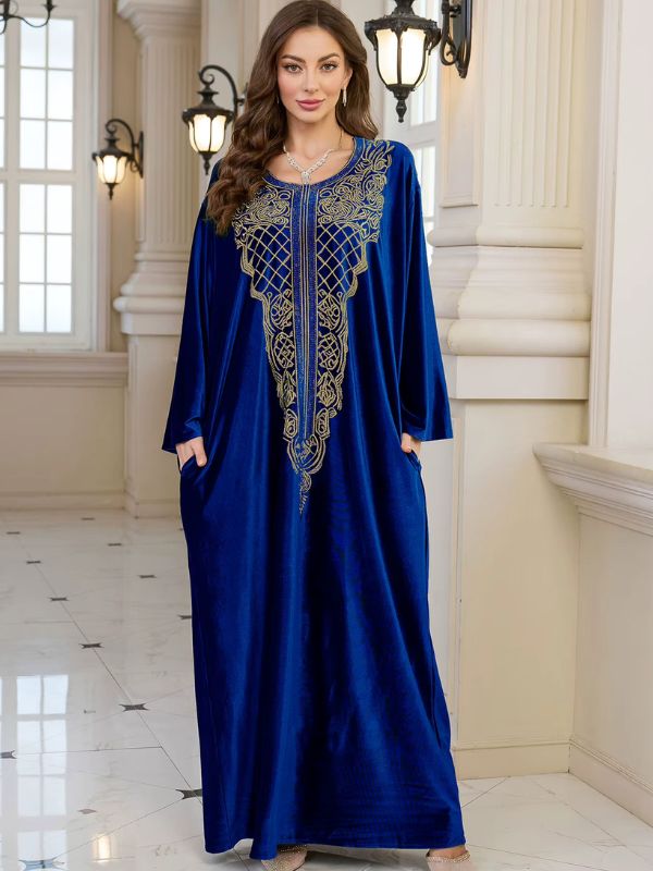 Abaya Dubaï en velours bleu roi, vue de face en pied, plastron brodé de fils dorés, mains dans les poches latérales et manches longues amples