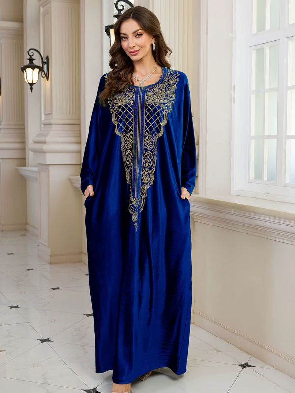 Abaya Dubaï en velours bleu roi, vue de face, mains glissées dans les poches latérales, broderie dorée sur le plastron