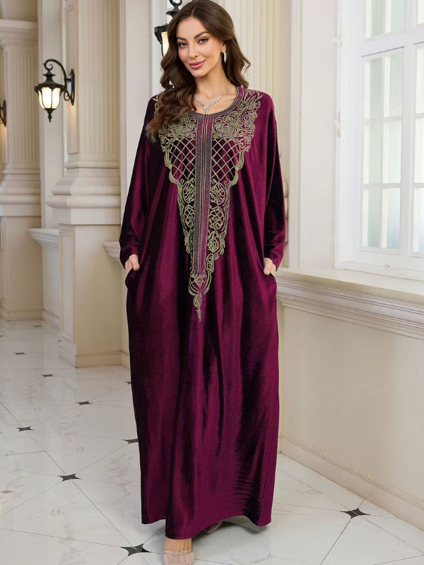Abaya Dubaï en velours bordeaux, vue de face, mains glissées dans les poches latérales, broderie dorée sur le plastron