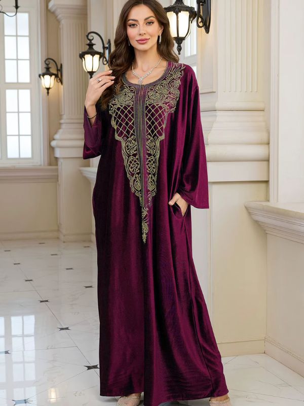 Abaya Dubaï en velours bordeaux, vue de trois quarts, une main dans la poche et l'autre relevée près de l'épaule, manches longues amples