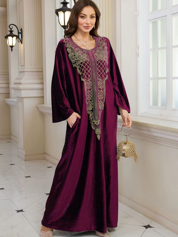 Abaya Dubaï en velours bordeaux, mannequin tenant un sac à main doré, robe ample et plastron brodé de fils dorés
