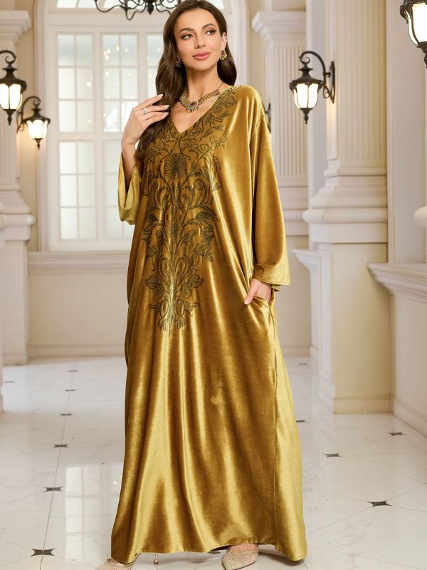 Abaya Dubaï en velours moutarde, vue de face, une main relevée près de l'oreille, l'autre dans la poche, broderie florale sur la poitrine