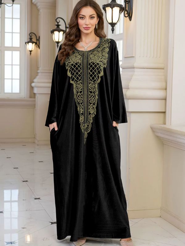 Abaya Dubaï en velours noir, vue de face en pied, plastron brodé de fils dorés, mains dans les poches latérales et manches longues amples