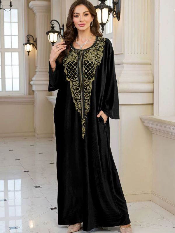 Abaya Dubaï en velours noir, vue de trois quarts, une main relevée près de l'épaule, l'autre dans la poche, plastron brodé doré