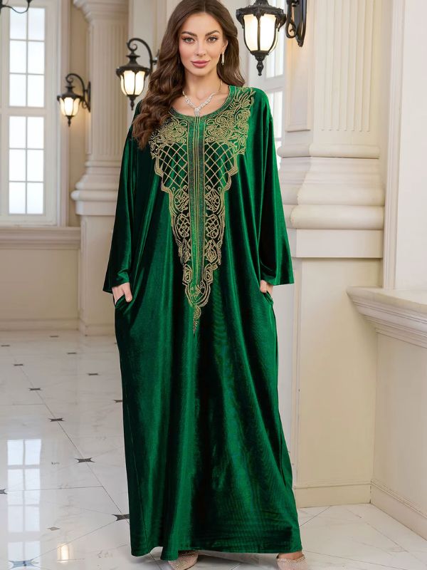 Abaya Dubaï en velours vert émeraude, vue de face en pied, plastron brodé de fils dorés, mains dans les poches latérales et manches longues amples