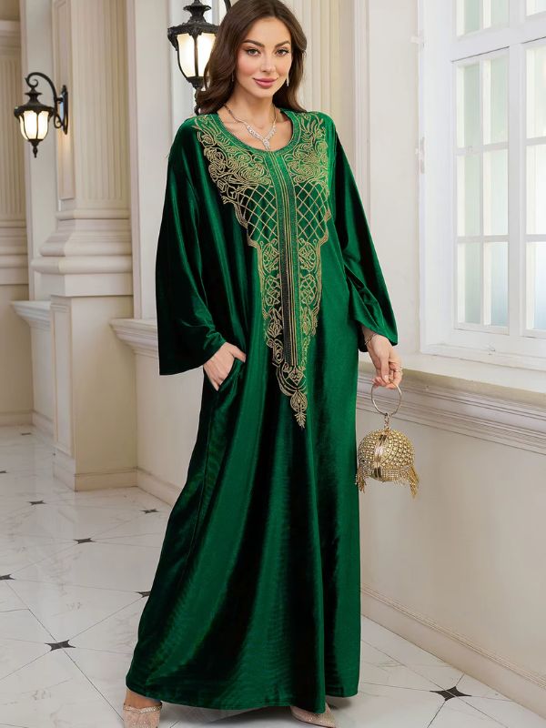 Abaya Dubaï en velours vert émeraude, mannequin tenant un sac à main doré, robe ample et plastron brodé de fils dorés
