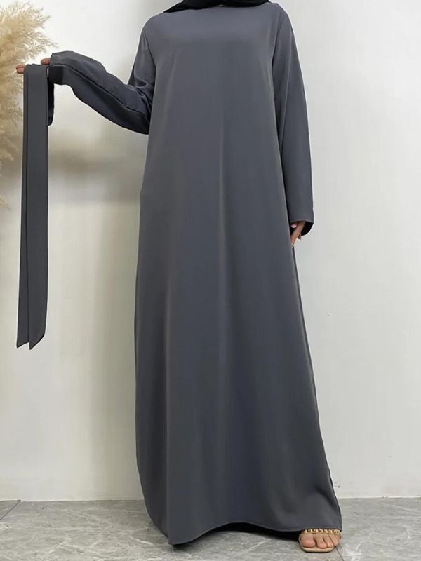 Une femme est debout, vêtue d'une abaya grise élégante. La robe est longue et fluide