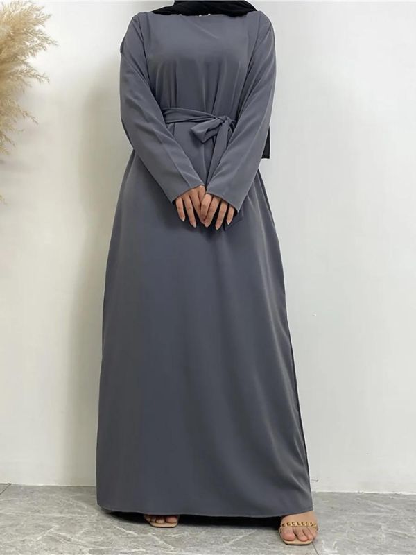 Une femme est debout, vêtue d'une abaya grise élégante. La robe est longue et fluide