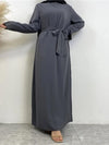 Une femme est debout, vêtue d&#39;une abaya grise élégante avec une ceinture nouée à la taille, ajoutant une touche de sophistication. Elle porte un hijab noir, créant un contraste classique avec la robe grise