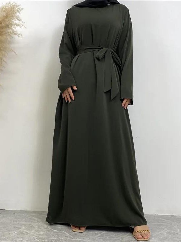 Une femme porte une abaya kaki, combinée avec un hijab noir. La tenue est fluide, couvrant entièrement le corps de manière élégante et modeste. Elle se tient droite, les bras légèrement écartés, mettant en valeur la longueur et la simplicité de la robe. Le fond neutre accentue l'élégance sobre de l'ensemble