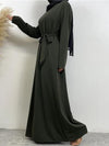 Vue de profil, la femme porte une abaya kaki ceinturée à la taille, avec un hijab noir couvrant la tête et tombant sur les épaules. Elle pose en tournant légèrement le corps, une main tenant le bout de l&#39;abaya. La tenue est élégante et fluide, mettant en valeur la simplicité et la sophistication du style. L&#39;arrière-plan reste neutre, permettant de se concentrer sur la tenue