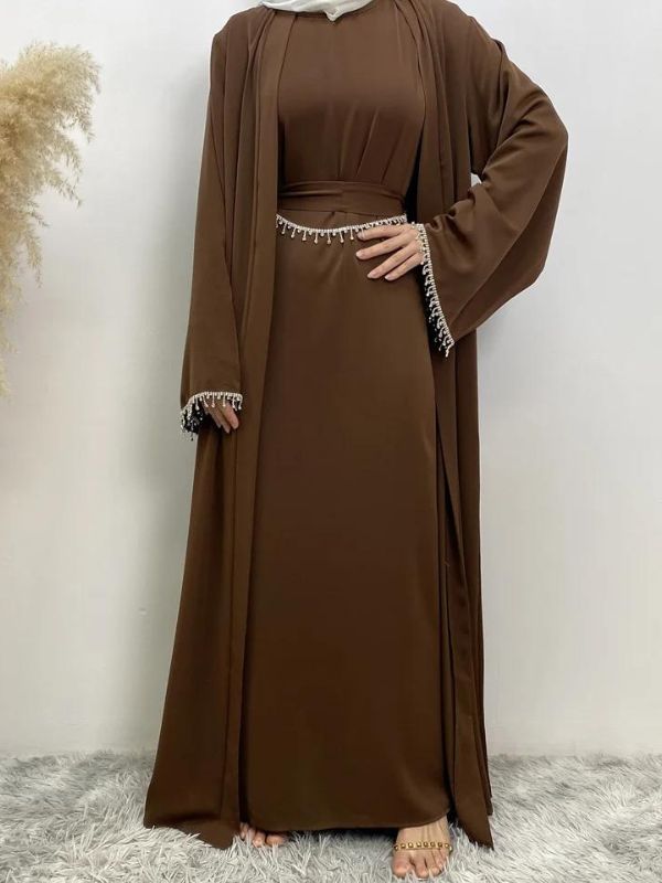 Femme portant une abaya marron avec des broderies blanches aux manches et à la taille, tenant une main sur la hanche, portant un hijab blanc, posant sur un fond neutre avec un vase de pampas à gauche