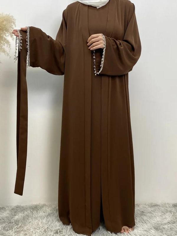 Femme portant une abaya marron avec des broderies blanches aux manches et à la taille, tenant une main sur la hanche, portant un hijab blanc, posant sur un fond neutre avec un vase de pampas à gauche