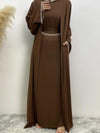 Femme en abaya marron avec des broderies blanches aux manches et à la taille, portant un hijab blanc, posant avec une main sur la poitrine et l&#39;autre dans une poche, sur un fond neutre avec un vase de pampas à gauche