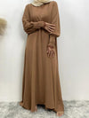 La femme porte une abaya marron clair, longue et fluide, qui descend jusqu&#39;au sol. La tenue est élégante et modeste, avec des manches longues plissées au niveau des poignets, ajoutant une touche de style et de sophistication. Elle porte également un hijab de couleur beige clair qui complète harmonieusement l&#39;ensemble. Le tissu de l&#39;abaya semble léger et confortable, parfait pour un look à la fois chic et discret