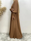 La femme est vue de profil, portant une abaya marron clair longue et fluide. Cette abaya met en valeur un style sobre et élégant avec des manches longues légèrement bouffantes, ornées de plis et de dentelle au niveau des poignets, ajoutant une touche délicate à l&#39;ensemble. Le hijab beige clair qu&#39;elle porte drape doucement autour de son cou, créant un contraste subtil avec la couleur marron clair de l&#39;abaya. Le tissu de la robe semble léger et fluide, offrant un mouvement gracieux lorsqu&#39;elle se déplace.