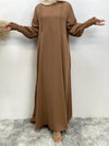 La femme porte une abaya marron clair qui est longue et fluide. Elle a les bras légèrement écartés, mettant en valeur les manches bouffantes de la robe, ornées de plis et de dentelle aux poignets. L&#39;abaya présente un design simple mais élégant, avec des détails discrets qui accentuent sa modestie et sa sophistication. Son hijab de couleur beige clair complète l&#39;ensemble avec douceur, ajoutant une touche harmonieuse au look global.