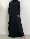 La femme porte une abaya longue de couleur noire. La robe est ample, descend jusqu&#39;au sol, et présente une coupe fluide et élégante. Elle est accompagnée d&#39;un hijab noir qui complète harmonieusement la tenue, recouvrant les épaules de manière soignée. La robe a des manches longues et une coupe droite, mettant en avant la simplicité et l&#39;élégance de l&#39;ensemble