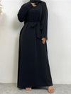 La femme porte une abaya noire longue et fluide qui descend jusqu&#39;au sol. La robe est simple et élégante, avec des manches longues et une coupe droite. Une ceinture noire est nouée autour de la taille, ajoutant une touche de définition à la silhouette. Elle porte également un hijab noir qui recouvre ses épaules, complétant harmonieusement la tenue. La simplicité et l&#39;élégance de l&#39;ensemble sont mises en valeur par le fond neutre, qui souligne les détails et la qualité de la tenue.