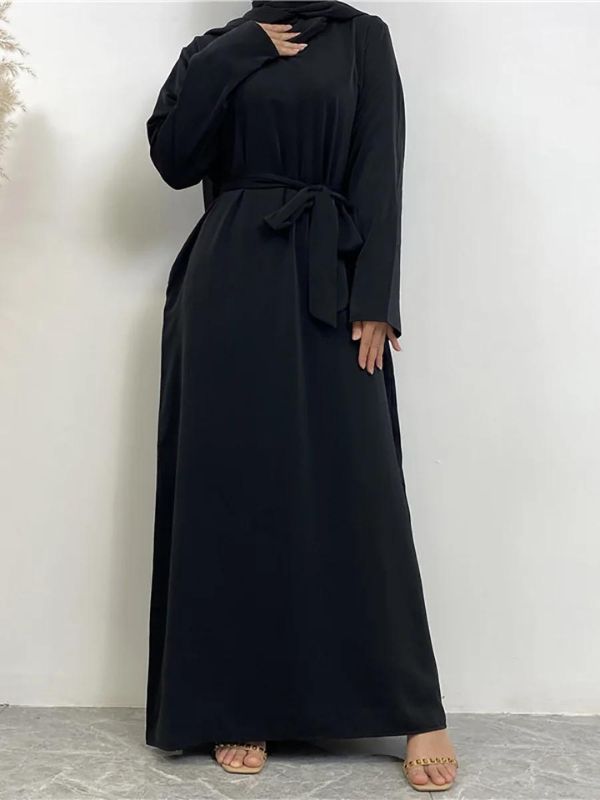 La femme porte une abaya longue de couleur noire. La robe est ample, descend jusqu'au sol, et présente une coupe fluide et élégante. Elle est accompagnée d'un hijab noir qui complète harmonieusement la tenue, recouvrant les épaules de manière soignée. La robe a des manches longues et une coupe droite, mettant en avant la simplicité et l'élégance de l'ensemble