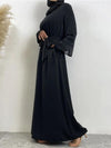 La femme porte une abaya noire longue et fluide, qui descend jusqu&#39;au sol. La robe est simple et élégante, avec des manches longues et une coupe droite. Une ceinture noire est nouée autour de la taille, ajoutant une touche de définition à la silhouette. Elle porte également un hijab noir qui recouvre ses épaules, complétant harmonieusement la tenue. La simplicité et l&#39;élégance de l&#39;ensemble sont mises en valeur par le fond neutre, qui souligne les détails et la qualité de la tenue