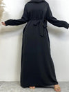  La femme porte une abaya noire fluide qui descend jusqu&#39;au sol, avec une ceinture noire nouée autour de la taille pour ajouter de la définition à la silhouette. La robe a des manches longues et une coupe droite, offrant une apparence simple mais élégante. Elle porte également un hijab noir qui complète harmonieusement la tenue.