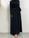 L&#39;image montre une femme portant une abaya noire vue de face. La robe longue et fluide atteint le sol, avec une ceinture noire nouée à la taille, créant une silhouette élégante et ajustée. La femme porte également un hijab noir, couvrant ses cheveux et ses épaules. La simplicité de la tenue est rehaussée par la qualité du tissu et la coupe soignée, offrant une apparence à la fois modeste et sophistiquée. Le fond neutre permet de bien mettre en valeur les détails de la tenue