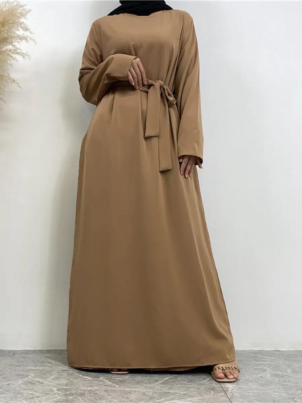 La femme est vue debout, portant une abaya nude ceinturée à la taille, créant une silhouette fluide et élégante. Elle porte également un hijab noir drapé, apportant un contraste à la tenue. Sa main droite est placée près de la ceinture, tandis que l'autre main repose sur le côté de son corps. L'arrière-plan est simple et minimaliste, mettant en valeur la tenue de la femme