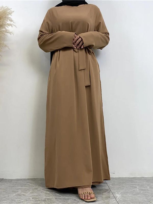 La femme est vue debout, portant une abaya nude ceinturée à la taille, créant une silhouette fluide et élégante. Elle porte également un hijab noir drapé, apportant un contraste à la tenue. Sa main droite est placée près de la ceinture, tandis que l'autre main repose sur le côté de son corps. L'arrière-plan est simple et minimaliste, mettant en valeur la tenue de la femme