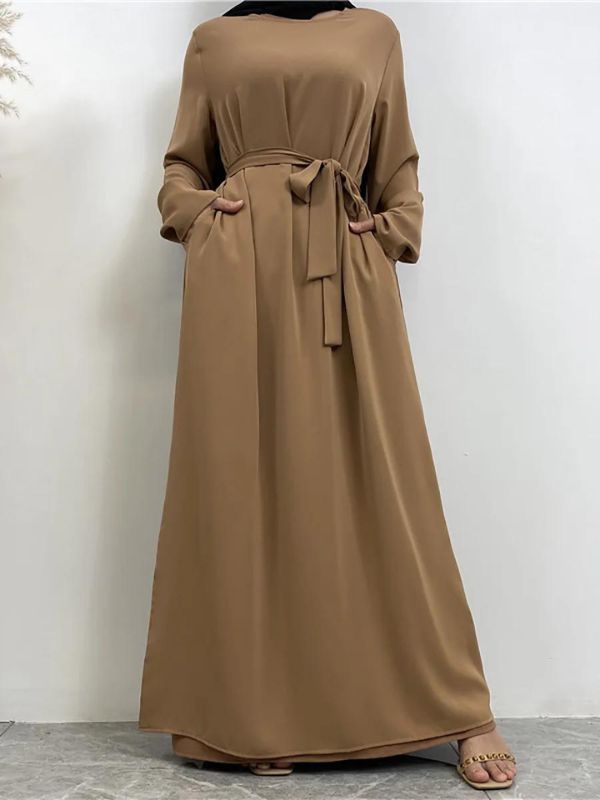 La femme porte une abaya nude ceinturée à la taille, avec un hijab noir. Elle se tient debout, les mains dans les poches, ajoutant une touche décontractée à son allure élégante. Le contraste entre le hijab noir et l&#39;abaya beige crée un look simple et raffiné. L&#39;arrière-plan neutre met en valeur les couleurs de sa tenue