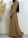 La femme porte une abaya nude, ceinturée à la taille, accompagnée d&#39;un hijab noir. Elle est vue de profil, ce qui met en valeur la fluidité et la simplicité élégante de la tenue. Le hijab noir contraste avec la couleur beige de l&#39;abaya, créant un look harmonieux. L&#39;arrière-plan neutre, avec une plante décorative à gauche, complète la composition visuelle