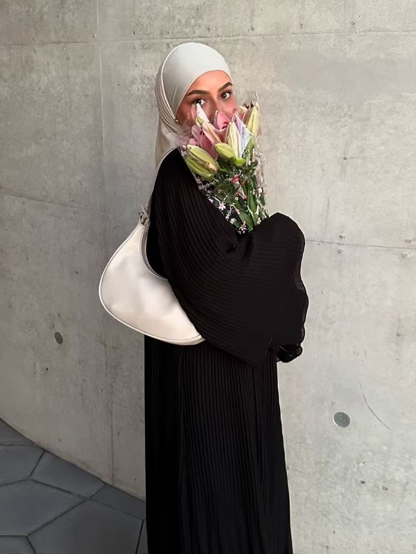 Femme de profil portant l'abaya plissée noire Lys d'Ébène à manches évasées, avec un hijab crème et un bouquet de lys à la main
