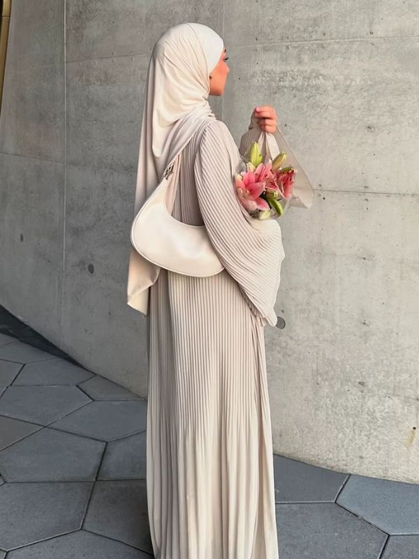 Abaya plissée beige crème Lys d'Ivoire vue de face, portée longue avec un hijab assorti, bras levé dévoilant la manche évasée