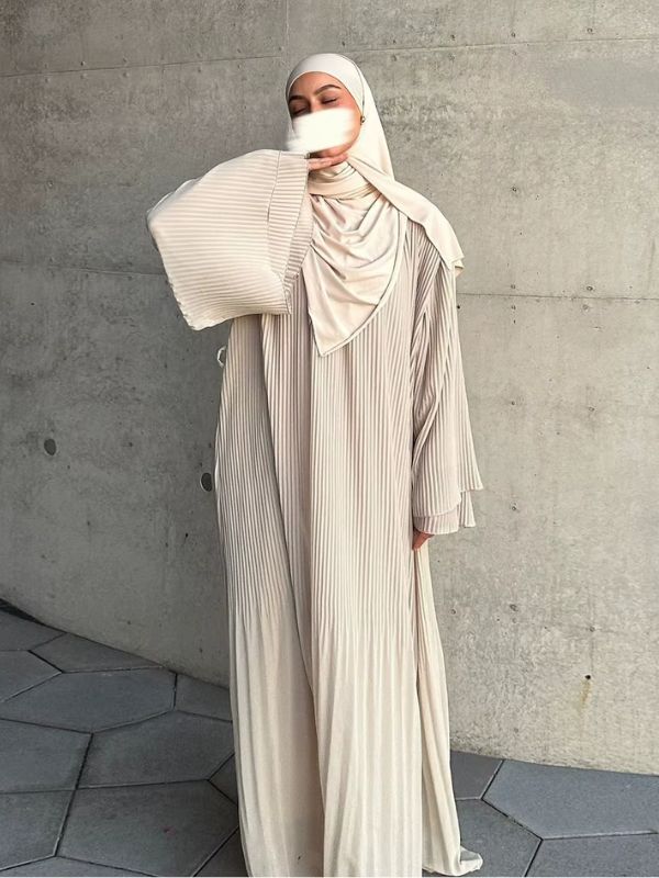 Abaya plissée beige crème Lys d'Ivoire vue de face, portée longue avec un hijab assorti, bras levé dévoilant la manche évasée