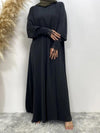 La femme porte une abaya noire en satin, qui lui confère un aspect élégant et fluide. Le tissu satiné ajoute une touche de luxe à la tenue, tandis que les manches longues sont ornées de dentelle délicate, ajoutant une dimension de sophistication supplémentaire. Elle complète son look avec un hijab assorti, légèrement différent en couleur, probablement dans une teinte de vert olive, créant un contraste subtil mais raffiné