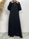 La femme porte une abaya noire en satin, caractérisée par une coupe fluide et élégante qui descend jusqu&#39;au sol. Le satin donne un aspect lustré et sophistiqué à la robe. Les manches sont longues et légèrement bouffantes, terminées par des poignets ornés de dentelle fine, ajoutant une touche féminine et délicate à la tenue. Elle tient le col de l&#39;abaya avec ses mains, ce qui met en valeur la fluidité du tissu et l&#39;élégance du design.