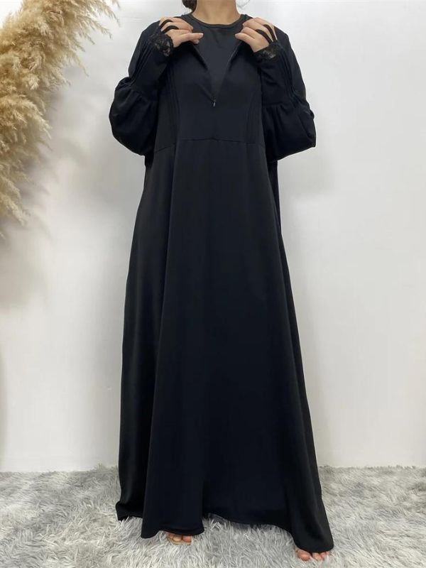 La femme porte une abaya noire en satin, qui lui confère un aspect élégant et fluide. Le tissu satiné ajoute une touche de luxe à la tenue, tandis que les manches longues sont ornées de dentelle délicate, ajoutant une dimension de sophistication supplémentaire. Elle complète son look avec un hijab assorti, légèrement différent en couleur, probablement dans une teinte de vert olive, créant un contraste subtil mais raffiné