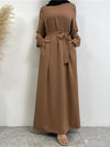 Une femme porte une abaya longue de couleur marron clair, ceinturée à la taille avec une ceinture assortie